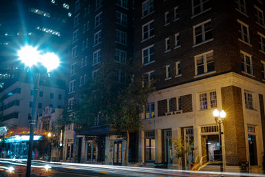 Nighttime downtown street scene with a historic brick building facade, glowing street lamps and long-exposure car light trails creating urban motion