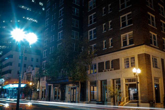 Nighttime downtown street scene with a historic brick building facade, glowing street lamps and long-exposure car light trails creating urban motion