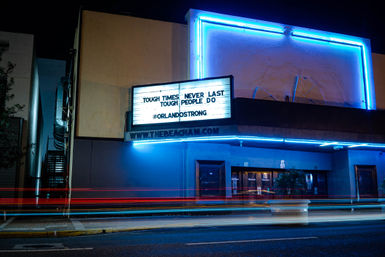 Nighttime street view of a neon-lit theater marquee in Orlando reading “Tough times never last, tough people do #OrlandoStrong,” blue neon outlining the building and colorful light trails from passing cars.