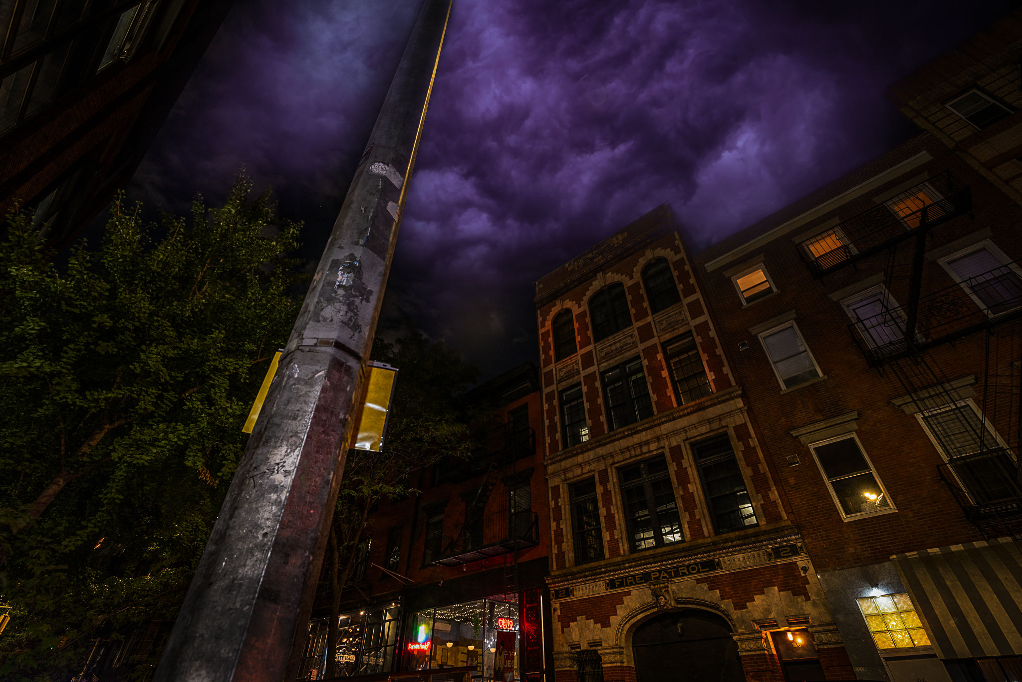 Nighttime urban street scene with a tall lamppost in the foreground, a historic brick building with fire escape and lit windows, neon cafe lights on the ground floor, and dramatic purple storm clouds overhead.