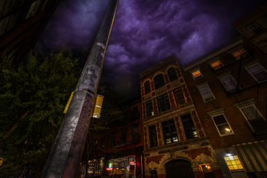 Nighttime urban street scene with a tall lamppost in the foreground, a historic brick building with fire escape and lit windows, neon cafe lights on the ground floor, and dramatic purple storm clouds overhead.