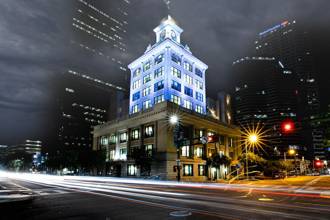 Long-exposure night view of an illuminated historic clock-tower building at a downtown intersection, with streaking car light trails and skyscrapers in the background.