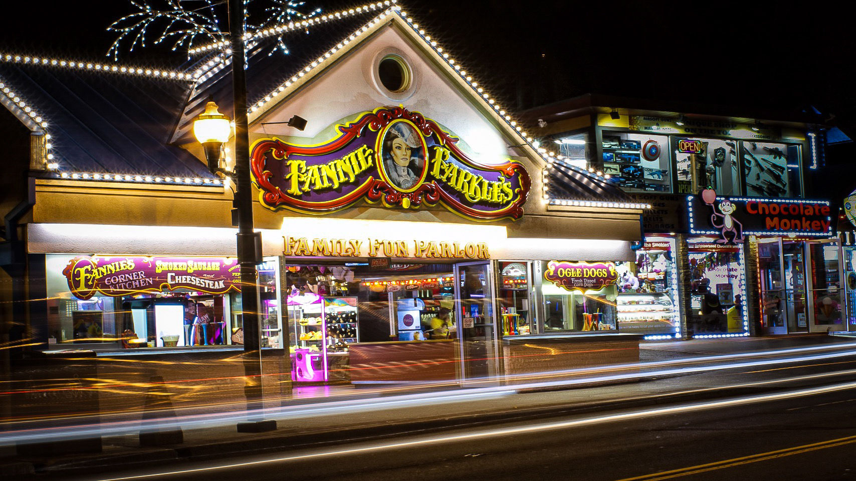Brightly lit family fun parlor and arcade at night with carnival-style signage, prize-filled windows, colorful neon lights and streaking car light trails on the street.