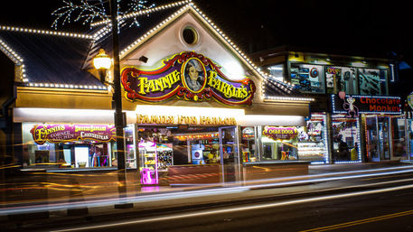 Brightly lit family fun parlor and arcade at night with carnival-style signage, prize-filled windows, colorful neon lights and streaking car light trails on the street.