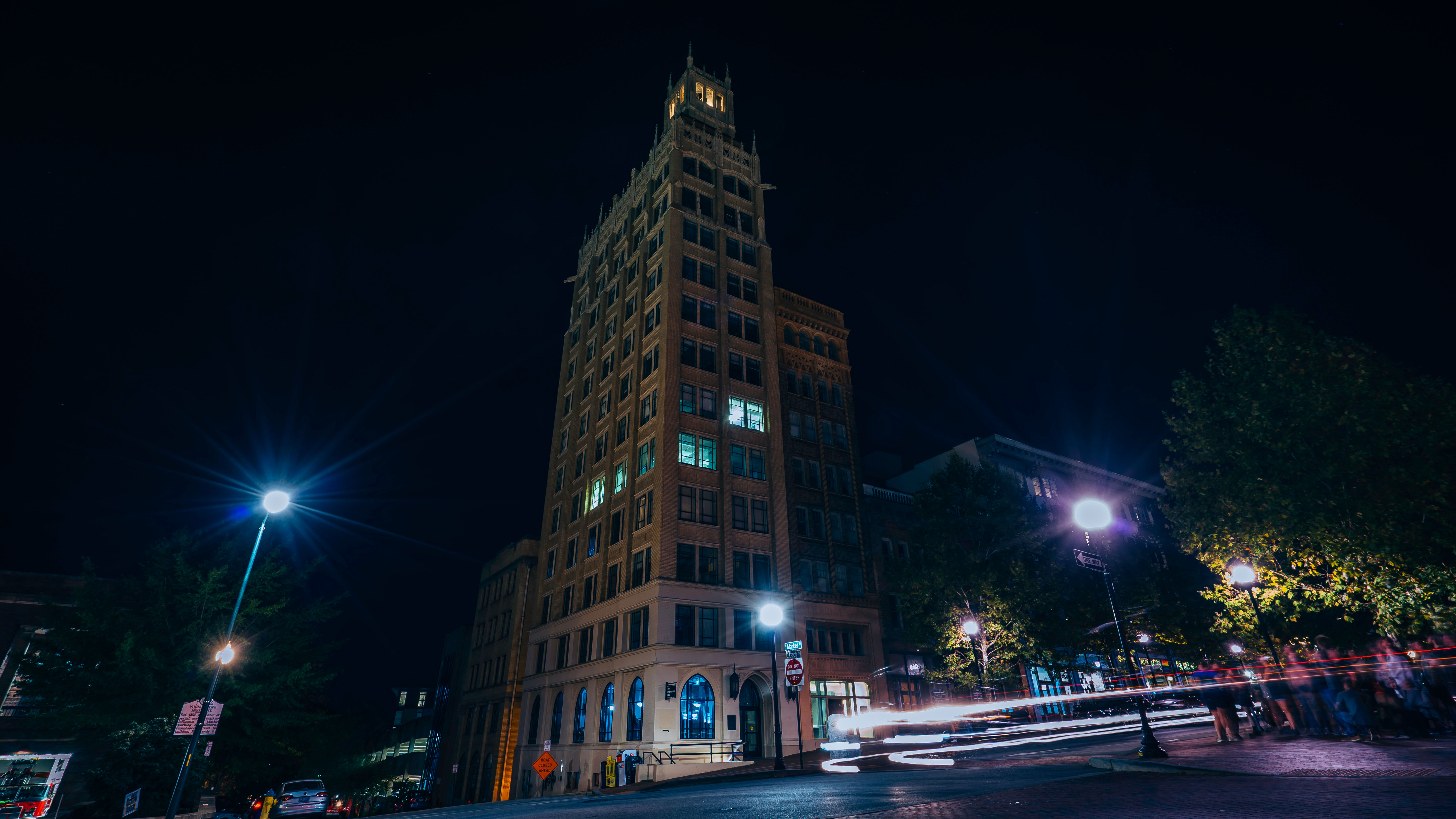 Night shot of a tall historic brick building in a downtown streetscape, glowing windows and street lamps with colorful car light trails streaking past.