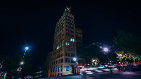 Night shot of a tall historic brick building in a downtown streetscape, glowing windows and street lamps with colorful car light trails streaking past.