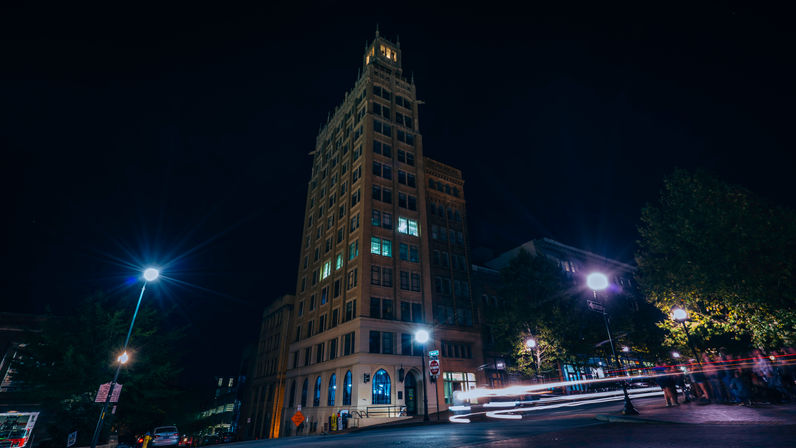 Night shot of a tall historic brick building in a downtown streetscape, glowing windows and street lamps with colorful car light trails streaking past.