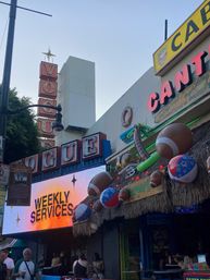 Vintage vertical theater marquee and bright LED sign reading "WEEKLY SERVICES" above a tropical-themed bar entrance on a bustling urban entertainment street, decorated with oversized inflatable footballs, beach balls, neon lights and a thatched awning.