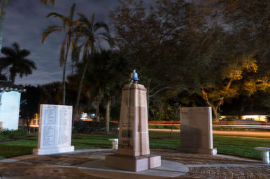 Nighttime view of a firefighter memorial — bell-topped stone obelisk with engraved name tablets on either side, palm trees overhead and streaking car light trails on a suburban street.
