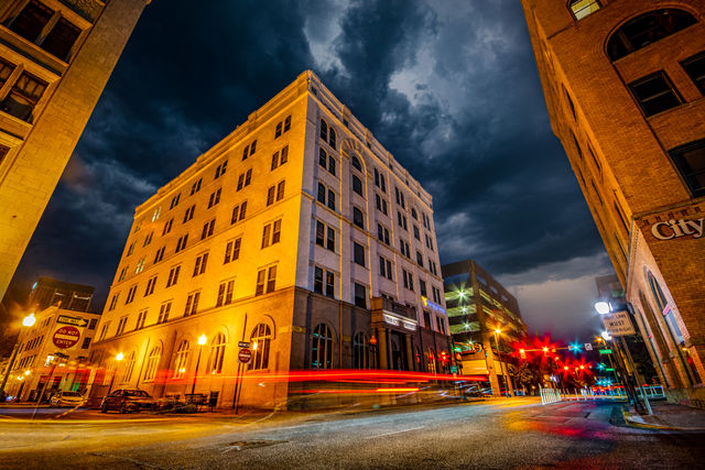 Nighttime downtown scene with a glowing historic corner building beneath dramatic storm clouds, streetlights and red car light trails streaking through a city intersection.