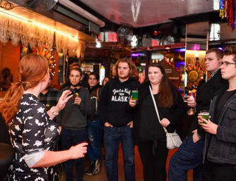 Red-haired host speaks to a small group of young adults holding bright green cocktails inside a cozy, colorful downtown bar with mirrored walls and festive lights.
