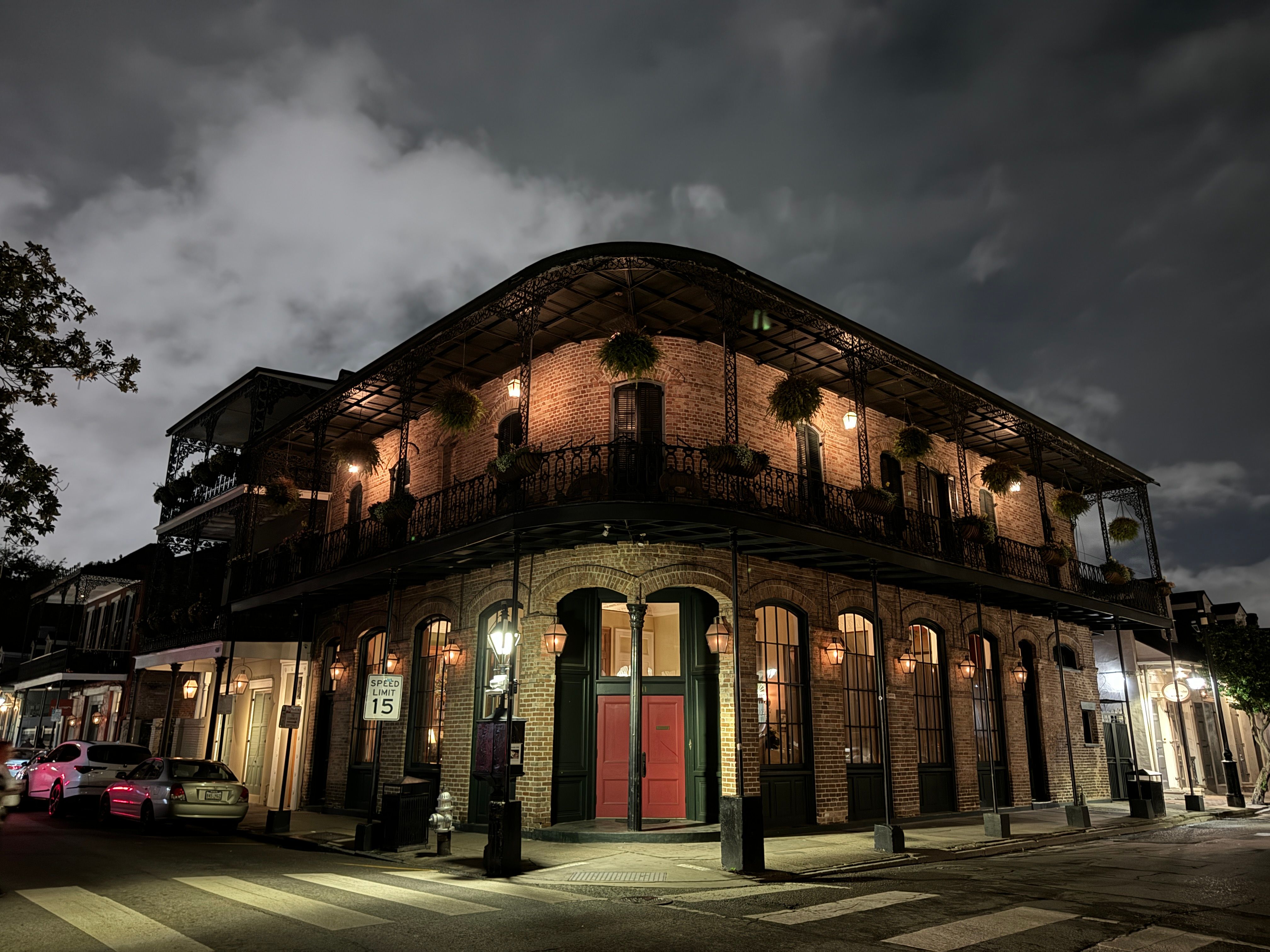 Historic brick corner building with ornate wrought-iron balcony and hanging plants in the New Orleans French Quarter at night, warm street lamps illuminating red doors and a quiet crosswalk.