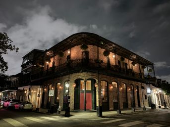 Historic brick corner building with ornate wrought-iron balcony and hanging plants in the New Orleans French Quarter at night, warm street lamps illuminating red doors and a quiet crosswalk.