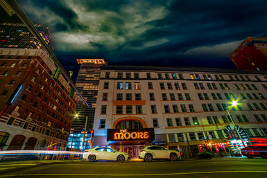 Vibrant nighttime downtown street with a glowing historic theater marquee, multi-story brick and stone buildings, parked cars and colorful light trails beneath dramatic stormy clouds.