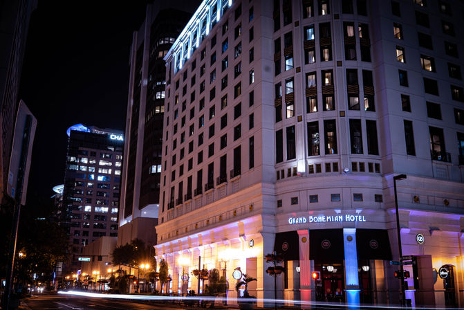 Downtown night scene — illuminated multi-story hotel facade with colorful uplighting, adjacent glass office towers, and light trails from passing cars.