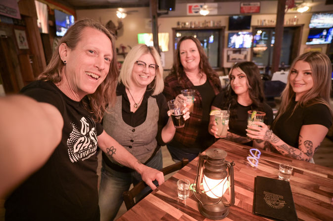 Five people smiling for a selfie at a rustic neighborhood bar, holding beers and cocktails around a wooden table with a glowing lantern — casual night out