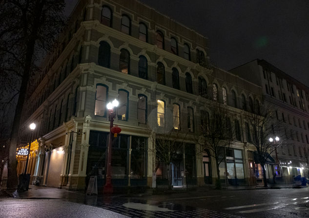 Historic multi-story downtown building with ornate arched windows at night, wet street reflecting glowing street lamps and a red lantern on a lamppost.