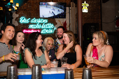 Lively group of friends laughing at a neon-lit bar counter, sipping drinks — one wearing a pink bachelorette sash — neon sign reading "Bad decisions make better stories" and a TV hockey game in the background, urban nightlife vibe.