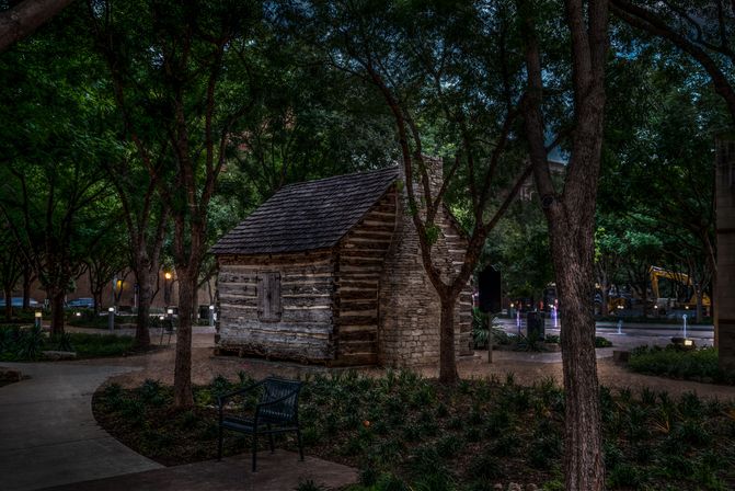 Historic log cabin with stone chimney in a shady downtown park at dusk, framed by oak trees, a bench, and illuminated fountains.
