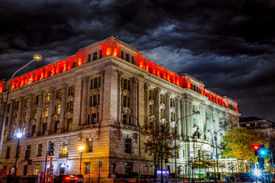 Historic neoclassical downtown building with red rooftop lighting at night beneath dramatic storm clouds, illuminated street lamps and moving traffic in the foreground.