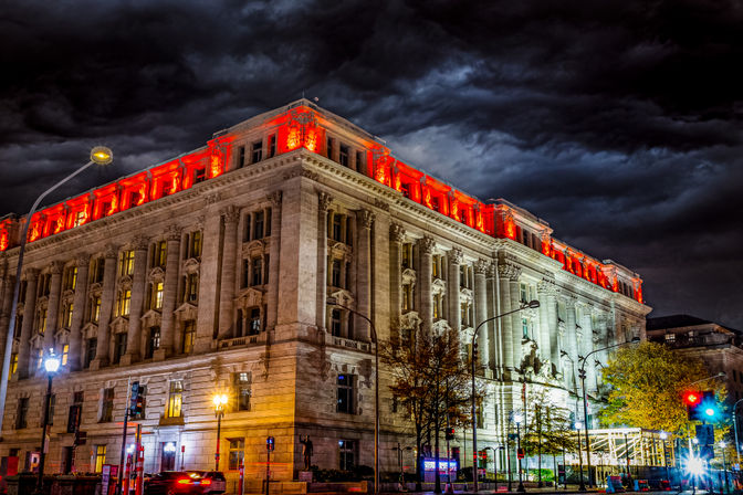 Historic neoclassical downtown building with red rooftop lighting at night beneath dramatic storm clouds, illuminated street lamps and moving traffic in the foreground.