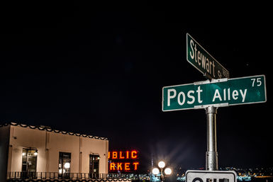 Nighttime waterfront scene with green street signs reading "Post Alley" and "Stewart St" in the foreground, neon red market letters and a lit building facade glowing above harbor lights.