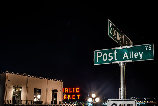 Nighttime waterfront scene with green street signs reading "Post Alley" and "Stewart St" in the foreground, neon red market letters and a lit building facade glowing above harbor lights.