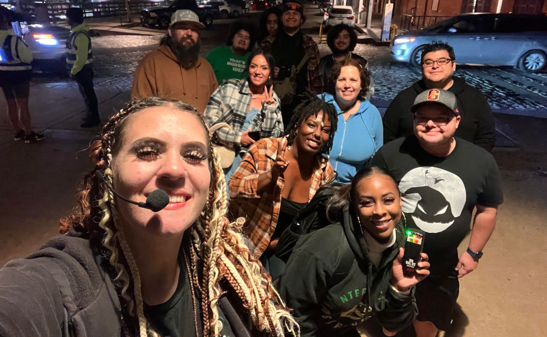 Group selfie of a smiling crowd on a cobblestone city street at night — front woman with braided hair and headset, friends behind flashing peace signs and holding a small device under streetlights.