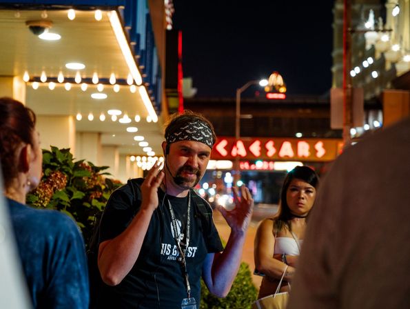 Bandana-wearing man animatedly gestures while speaking to a small group under marquee lights on a lively city sidewalk at night, with neon casino-style signage and illuminated buildings in the background.