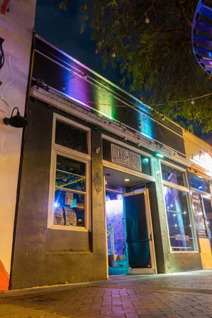 Vibrant downtown bar entrance at night — open doorway, rainbow neon wash, hanging string bulbs and sidewalk