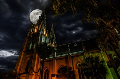 Gothic-style church with twin spires lit orange-green under a full moon and dramatic storm clouds, palm trees and hanging Spanish moss in the foreground.