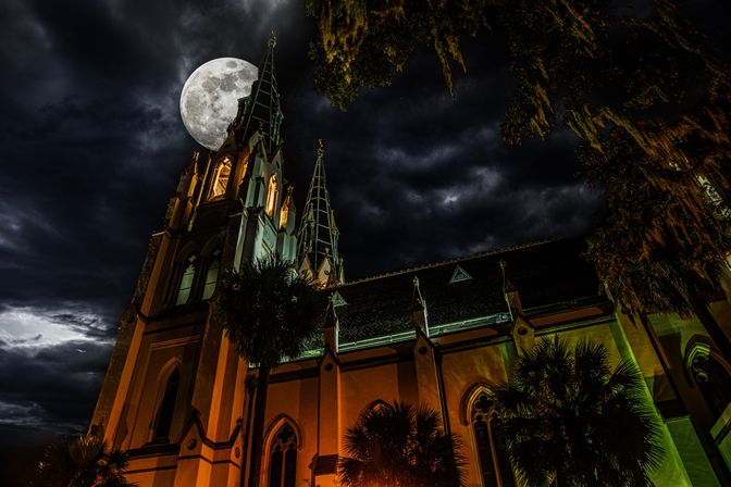 Gothic-style church with twin spires lit orange-green under a full moon and dramatic storm clouds, palm trees and hanging Spanish moss in the foreground.