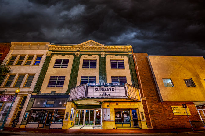 Historic three-story theater-style building on a downtown street with an illuminated marquee reading SUNDAYS AT 11AM, glowing glass doors, and dramatic storm clouds overhead.