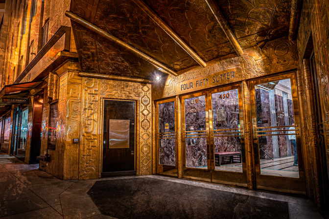 Golden Art Deco building entrance with ornate embossed facade, glass doors marked 450 revealing a marble lobby, nighttime downtown sidewalk scene