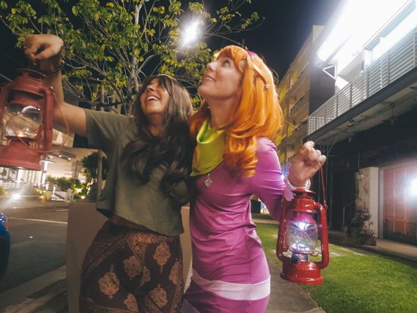 Two friends in costumes holding red lanterns on a city sidewalk at night, one in a purple dress with an orange wig and green scarf and the other in a green top and patterned skirt, laughing under streetlights.