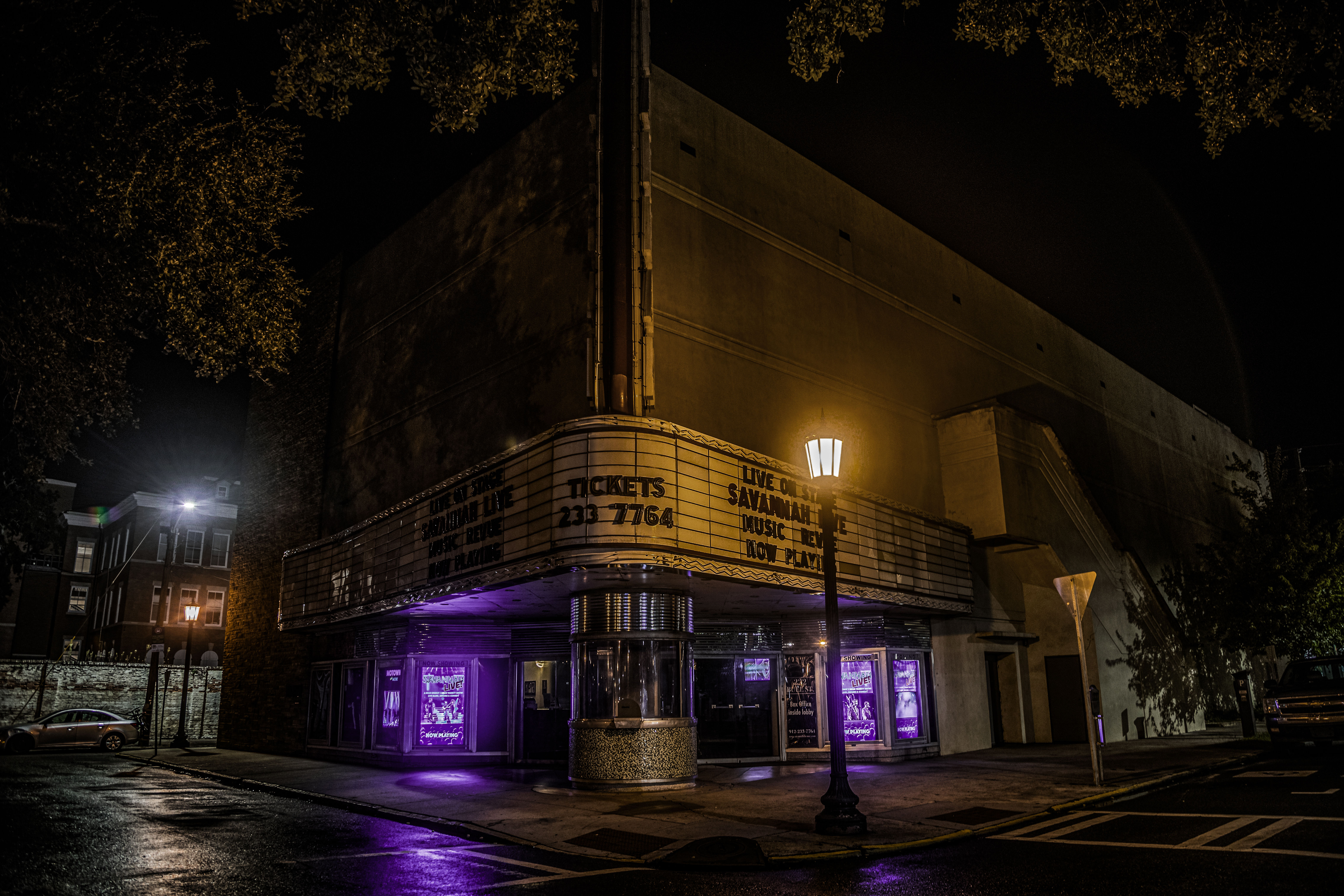Historic downtown theater at night with a glowing marquee and corner ticket booth bathed in purple light, wet sidewalk and streetlamp adding moody reflections.