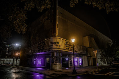 Historic downtown theater at night with a glowing marquee and corner ticket booth bathed in purple light, wet sidewalk and streetlamp adding moody reflections.