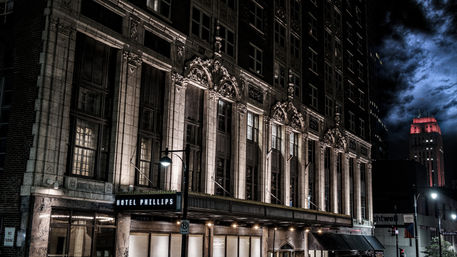 Historic urban hotel facade at night with ornate stone columns and illuminated windows over a lit entrance, moody cloudy sky and a distant red-lit tower on a city street.