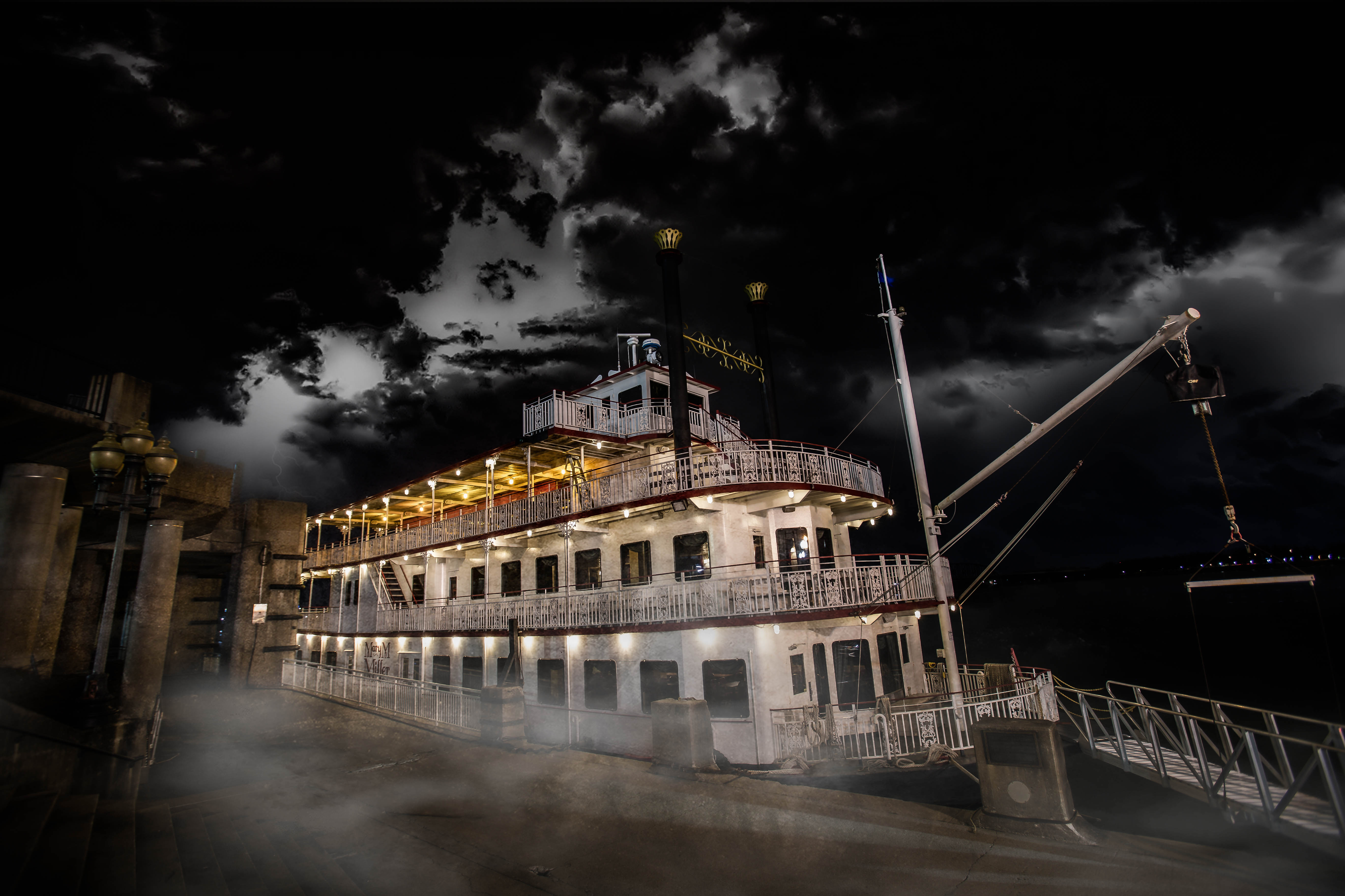 Moody riverboat docked at a riverside pier at night, glowing deck lights, swirling mist along the walkway and dramatic storm clouds overhead