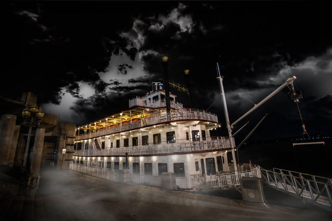 Moody riverboat docked at a riverside pier at night, glowing deck lights, swirling mist along the walkway and dramatic storm clouds overhead