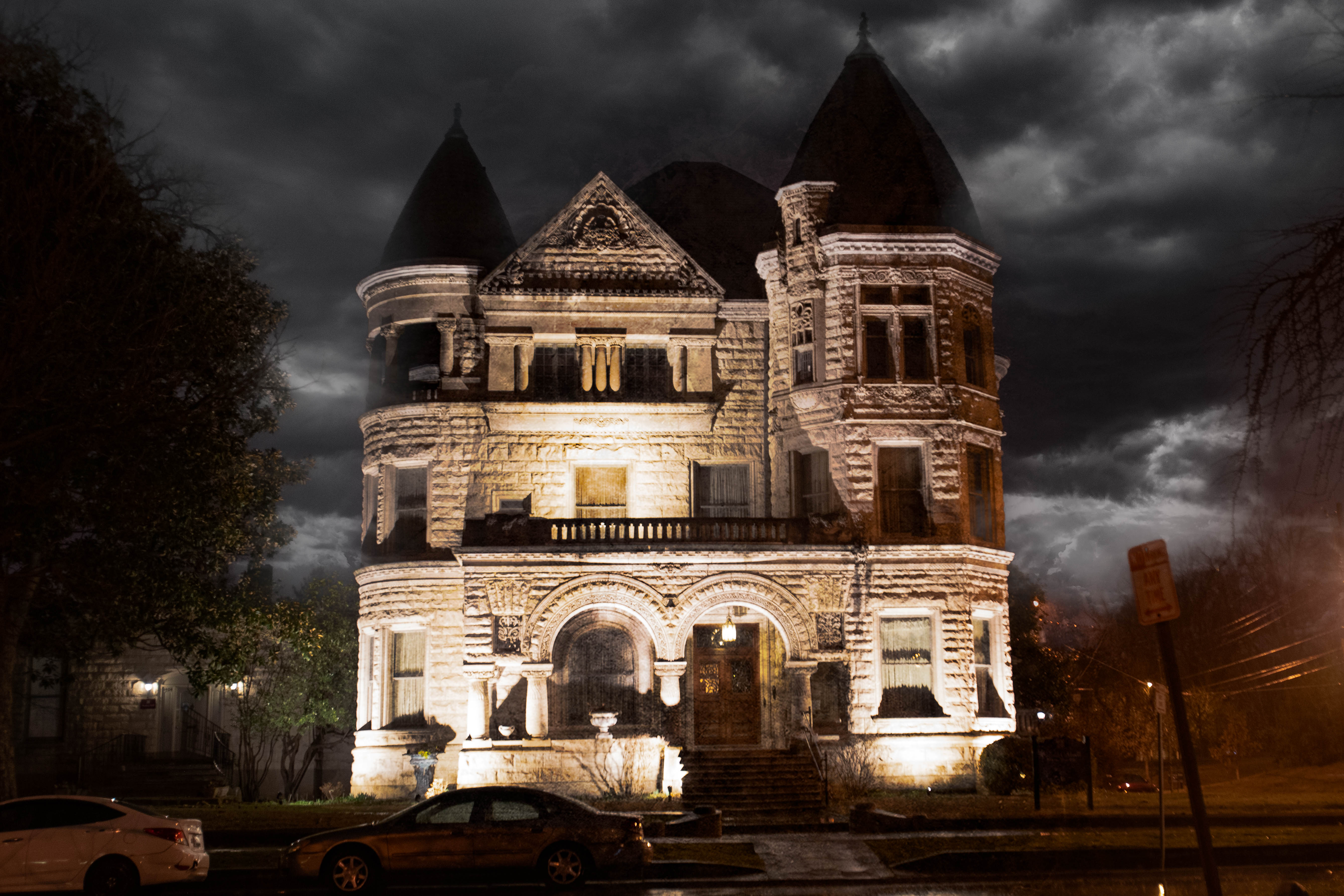 Moody, castle-like Victorian mansion at night — illuminated stone façade with turrets and arched entrance under dramatic stormy clouds, parked cars along a wet street.