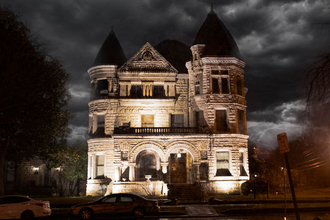 Moody, castle-like Victorian mansion at night — illuminated stone façade with turrets and arched entrance under dramatic stormy clouds, parked cars along a wet street.