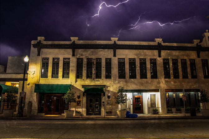 Two-story historic downtown building with green awnings and lit entrances on an empty city street at night, dramatic purple sky with lightning bolts