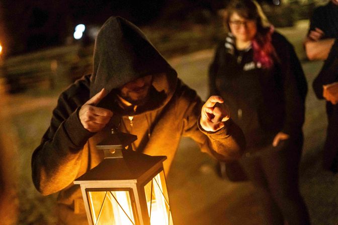 Outdoor nighttime scene with a hooded person leaning over a glowing lantern, warm light illuminating their hands and face while a small group watches blurred in the background.