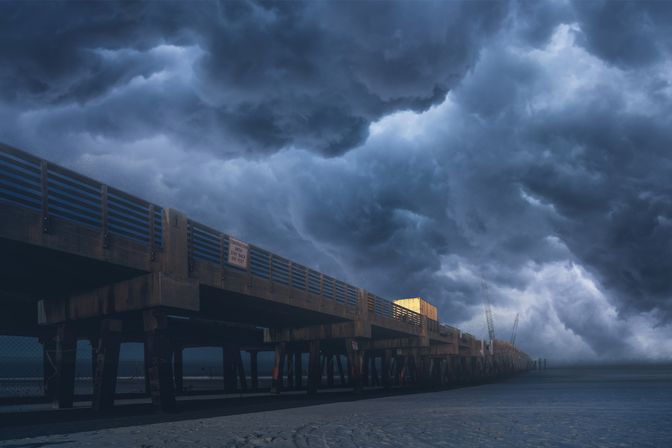 Cinematic storm clouds rolling over a long wooden coastal pier on a sandy beach, dark ocean and distant cranes under a brooding sky