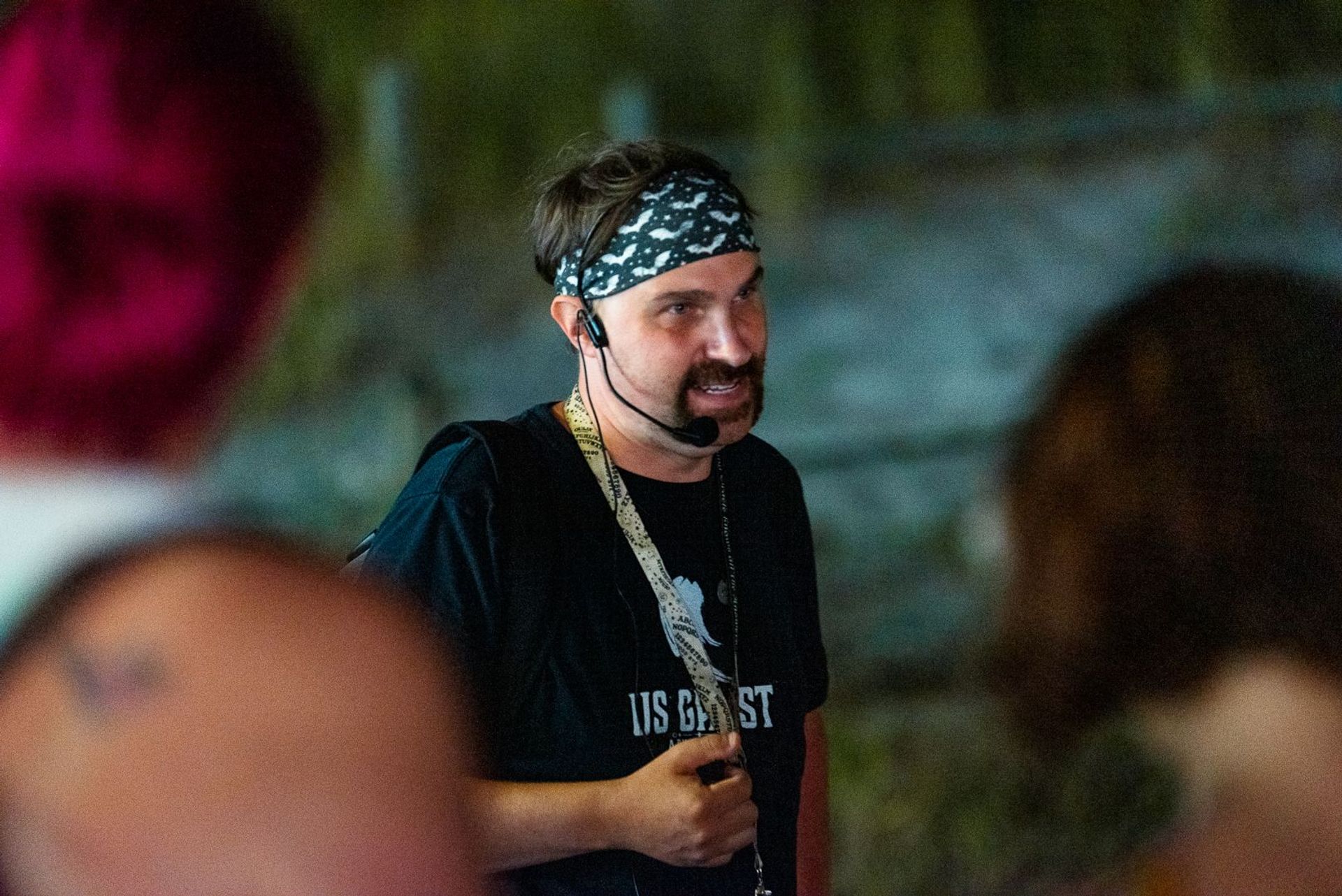 Animated tour guide wearing a patterned bandana and headset microphone speaking to a small group during an outdoor night walking tour, black t-shirt and lanyard visible.