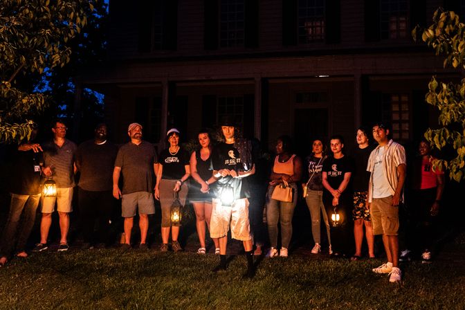 Group of adults on a nighttime ghost tour standing on a lawn in front of a large historic house, holding glowing lanterns and flashlights