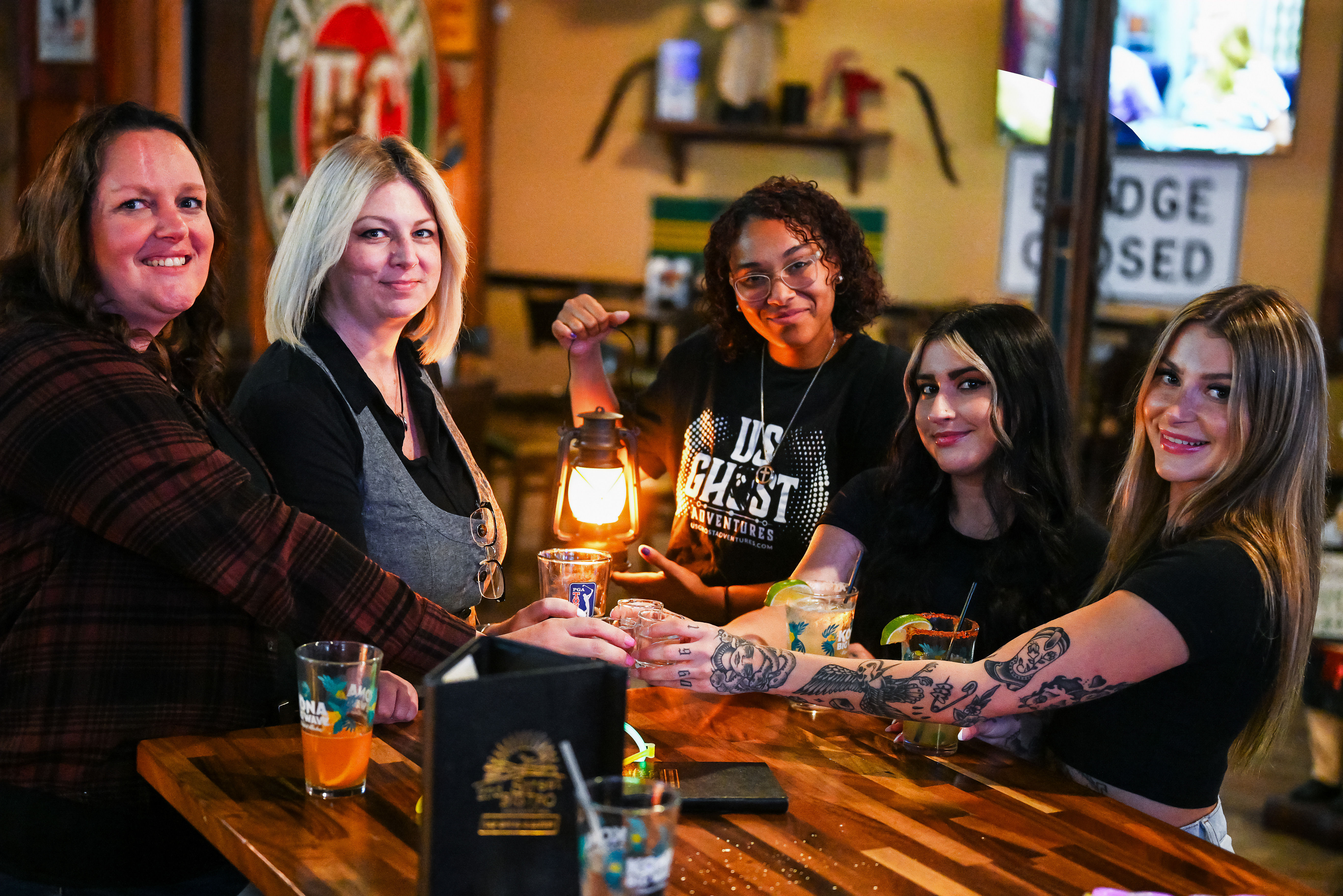 Five women smiling at a wooden bar, raising cocktails and a glowing lantern for a toast, visible arm tattoos and colorful drinks in a cozy pub setting.