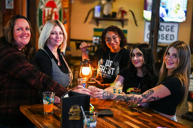 Five women smiling at a wooden bar, raising cocktails and a glowing lantern for a toast, visible arm tattoos and colorful drinks in a cozy pub setting.