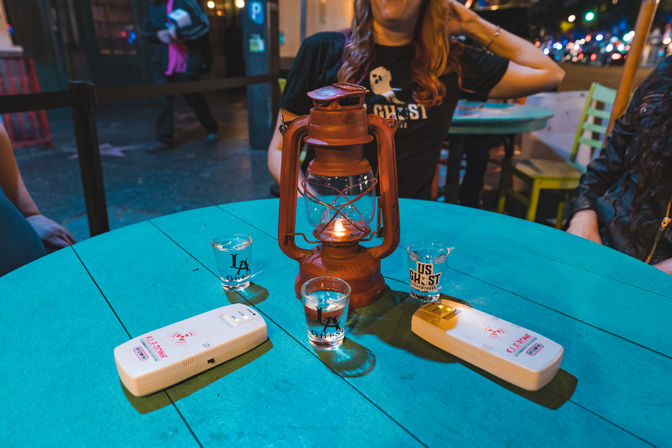 Nighttime outdoor patio scene: teal round table with a lit vintage lantern centerpiece, three shot glasses with logos and two white pager buzzers, blurred city street and seated people in the background.
